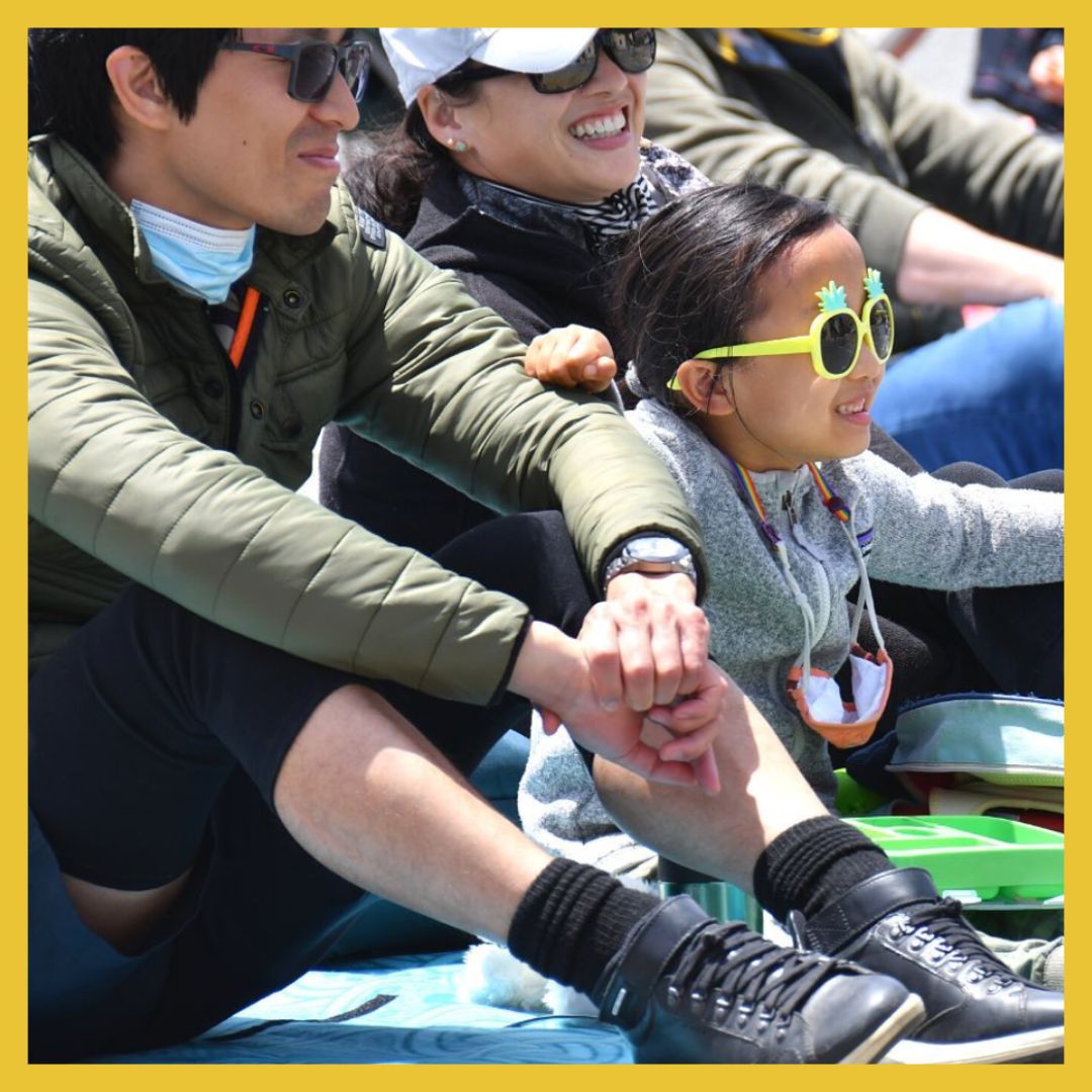 Image Description: A smiling family wearing sunglasses sits on blankets outside and watches a performance outside of the frame.