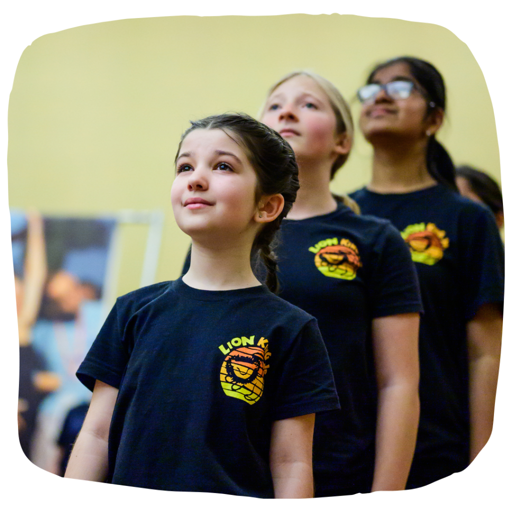 Young girls are in a line all wearing a tee shirt that has a lionk king jr. logo on it. They look up in aspiration.