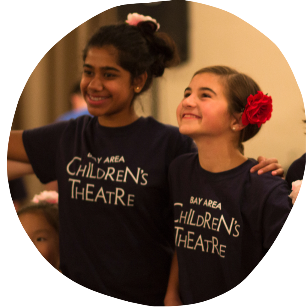 Two youth (both wearing Bay Area Children's Theatre Tee-Shirts) smile with flowers in their hair.