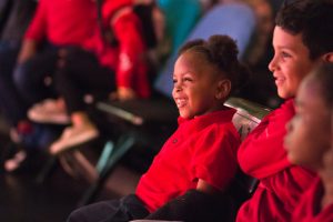 Children wearing the same red polo shirt, sit in an audience smiling.
