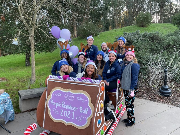 A photo of the Bay Area Children's Theatre Staff in 2021, posing in Santa's Sleigh. The sign on the sleigh says "Purple Reindeer Ball 2021".