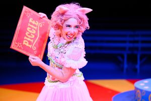 Piggie (played by Bri Reads) is dressed in a pink dress, pink wig and pink ears and holds a large envelope that says "Piggie" on it. Piggie smiles while leaning and hugging the envelope.