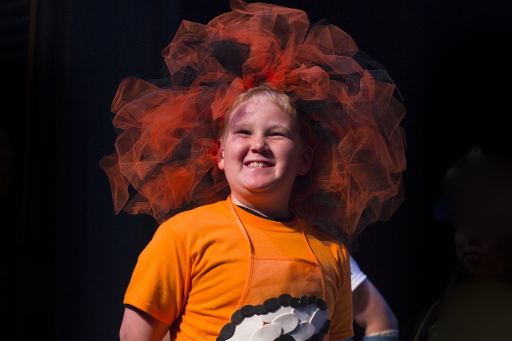 Child in an orange shirt and orange poofy crown smiles as he stands on stage