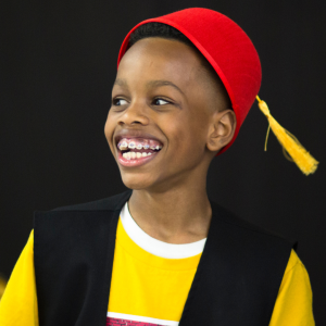 11 year old wearing a red hat with a yellow tassel hanging from the top, a black vest and yellow tee-shirt, smiles widely. He has braces and has his head slightly angled to the left